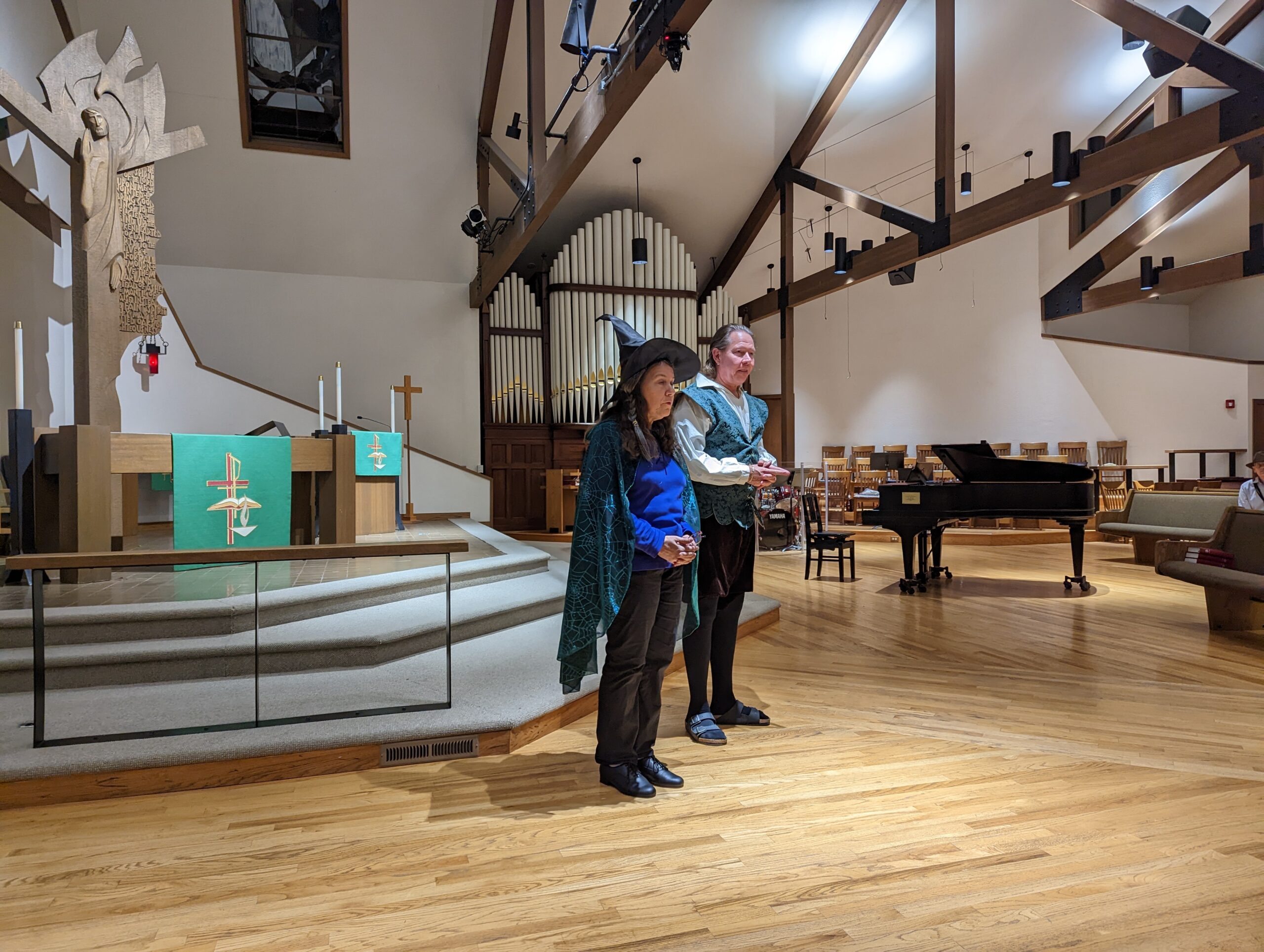 Woman and man in costume stand in front of audience with organ and piano in background