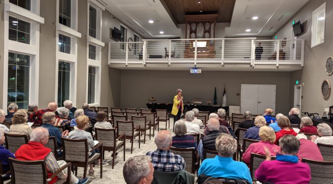 View of organ in gallery along with audience and woman speaking