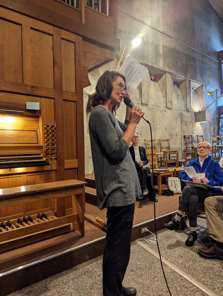 woman with microphone stands in front of organ addressing audience