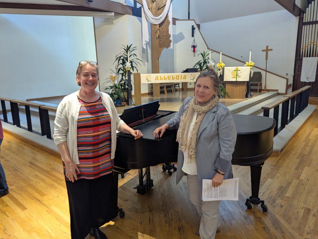 Two women stand in front of a grand piano in a church